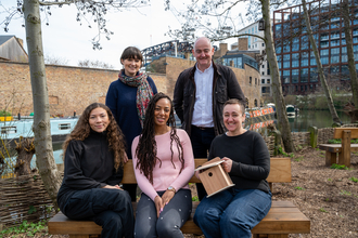 A woman and a man stood behind three women sat on a bench.